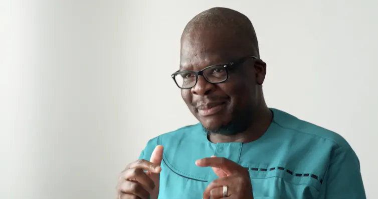 A bald African man in horn-rimmed glasses smiles and holds his hands up expressively as he talks, wearing a green African shirt.