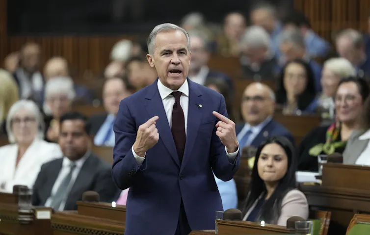Un hombre delgado con cabello corto y oscuro se señala el pecho con el dedo índice mientras habla.