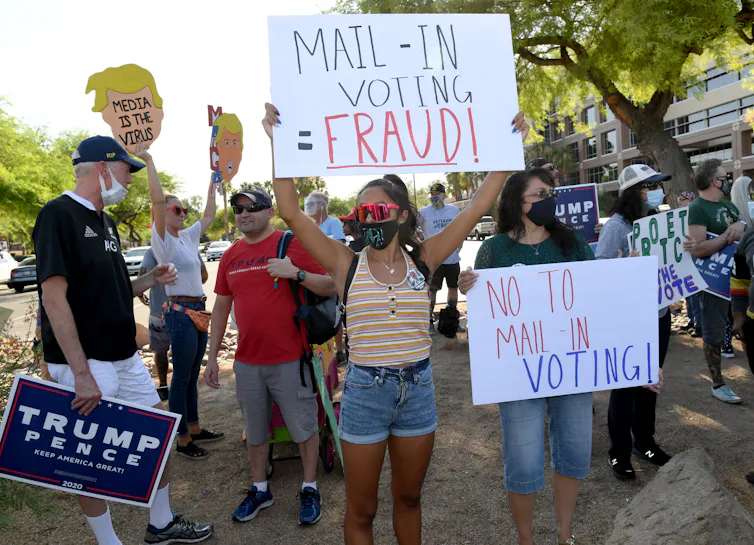 A group of protesters holding signs about mail-in voting fraud, outside a large building.