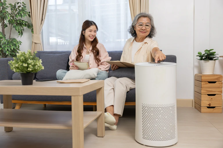 An older woman presses a button on an air purifier while sitting on a couch with a younger woman.