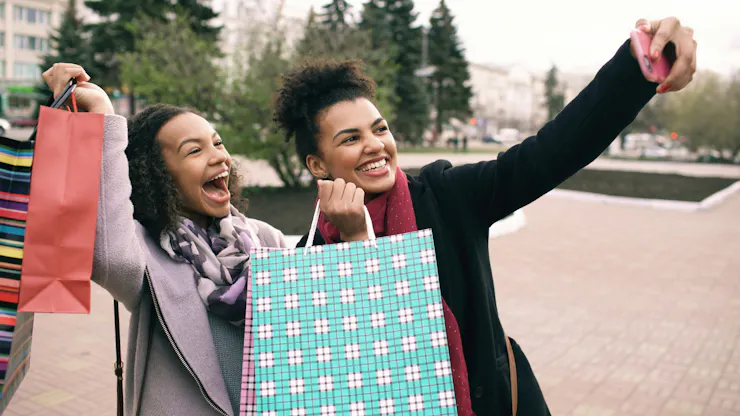 Two young Black women with shopping bags smile and laugh as they take a selfie after a mall sale.