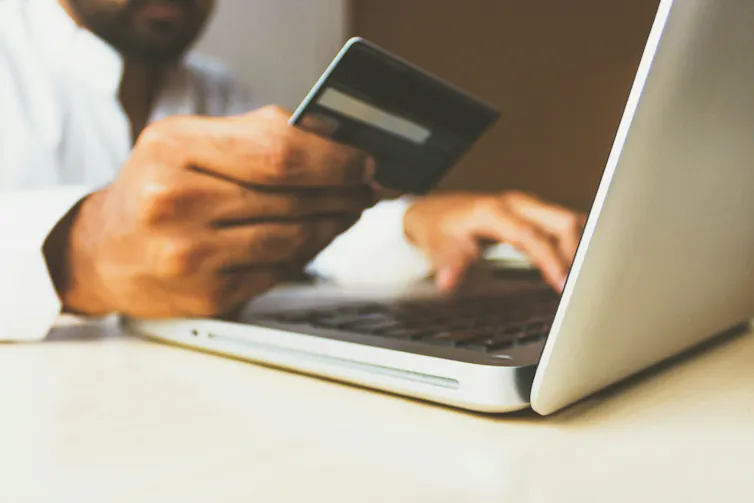 A man seated in front of a laptop holds a credit card in one hand while making an online purchase with the other.