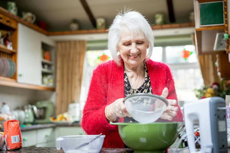 Older woman in kitchen sifting flour into green bowl.