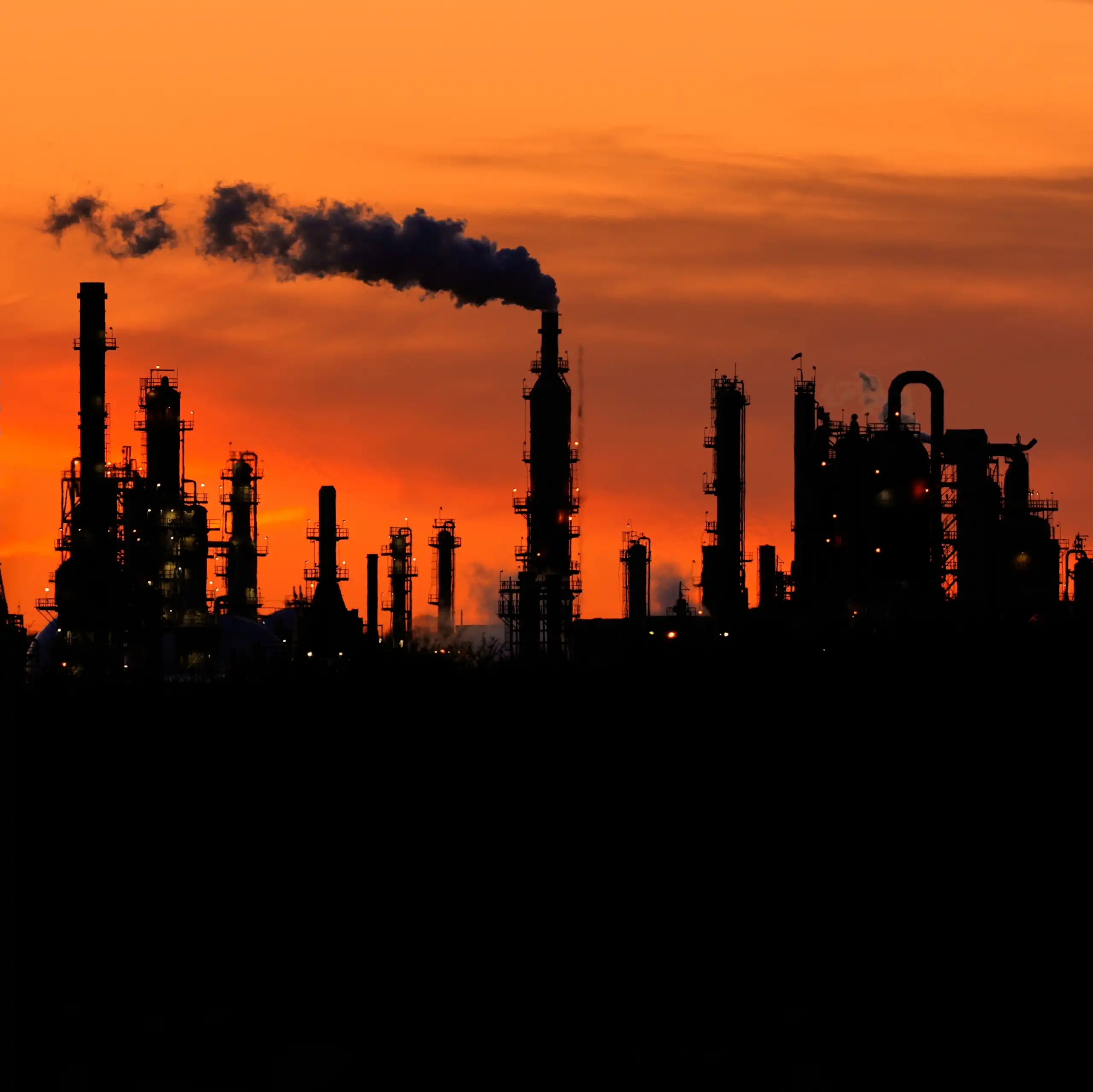 smoke stacks at an industrial plant silhouetted by an orange sky at sunset