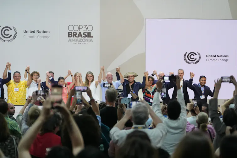 Esto es lo que se puede esperar de la primera conferencia sobre la transición a los combustibles fósiles 1 Las personas en una sala grande se paran juntas y levantan la mano, otras miran.