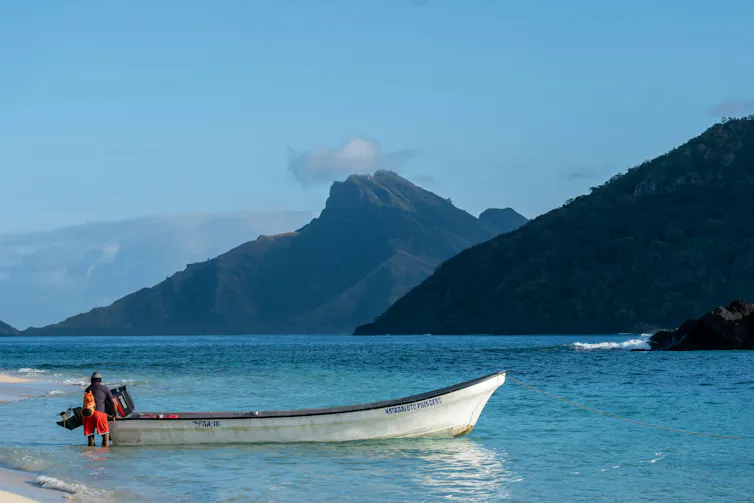 man standing next to banana boat in turquoise blue waters, mountains as backdrop.