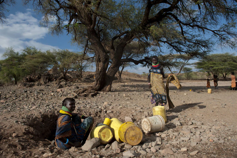 Woman seating on rocky, dry ground in a shallow hole surrounded by yellow jerry cans, while another woman stands nearby