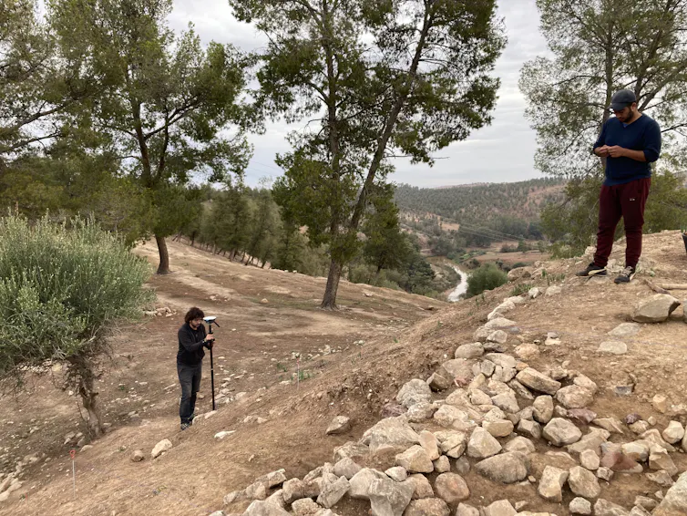 A man stands with a device on a tripod pointed at stacks of stones forming a ridge in the ground, another man standing above it, both dressed in black.