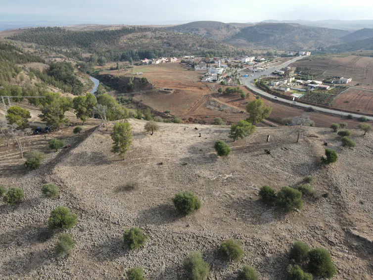 An aerial photograph of a sparse rocky outcrop with raised ridges at the top, a road and village in the distance.
