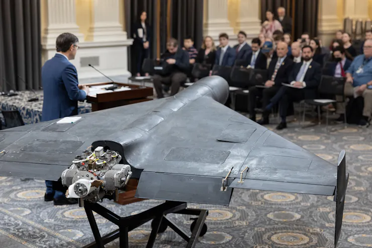 A man in a suit stands next to a drone and speaks to a group of seated people.