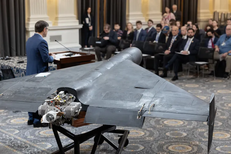 A man in a suit stands next to a drone and speaks to a group of seated people.