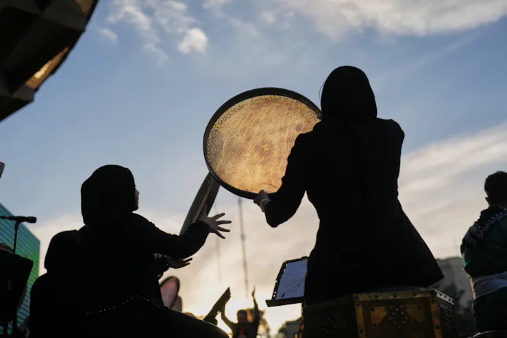 Two women, seen from behind, hold up a large flat drum against a cloudy blue sky.