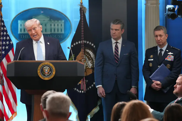 A man in a suit stands at a lectern next to an American flag while two younger men stand to the right.