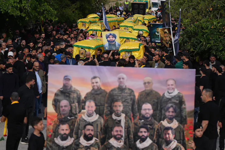 A crowd of people, many of them wearing black, walks holding coffins draped in yellow fabric and a large photo of 15 men in military uniforms.