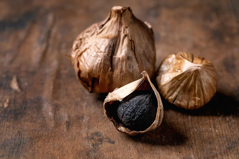 A whole bulb of black fermented garlic and both a peeled and unpeeled clove on a dark wooden table.