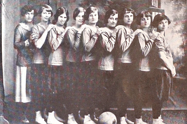 A black and white photo shows a row of teenage girls with their hands on each other's shoulders, all standing in a row and facing toward the right.