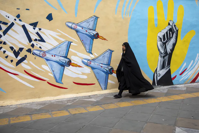 A woman in traditional Muslim garb walks past a wall with paintings of drones and a fist on it.