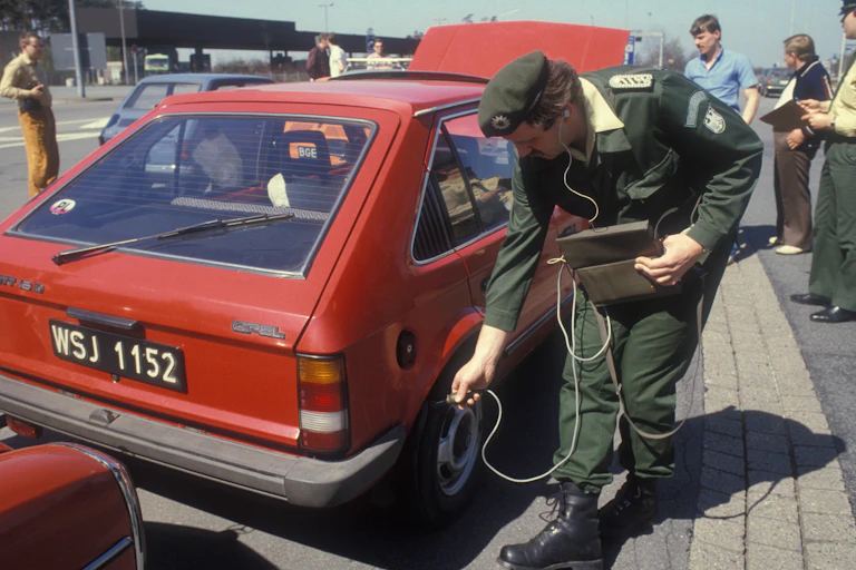 A man with a machine and wires stands next to an old red car.
