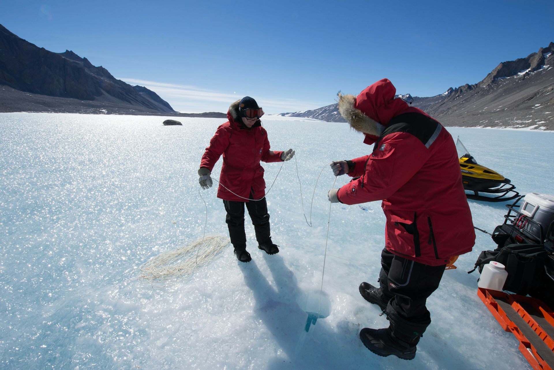 Dos hombres con abrigos rojos de nieve en un lago congelado con un cielo azul profundo en el fondo.