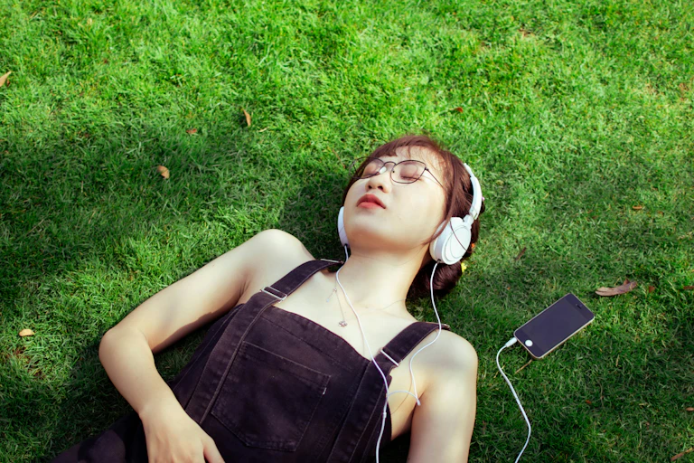 A woman lying on the grass in sunshine, listening to music through headphones.