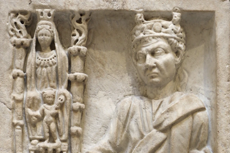 A marble relief with several figures engraved on it: on the side is a column with an adorned woman with another companion. Next to them is a man with a crown, with one hand on a pedestal.