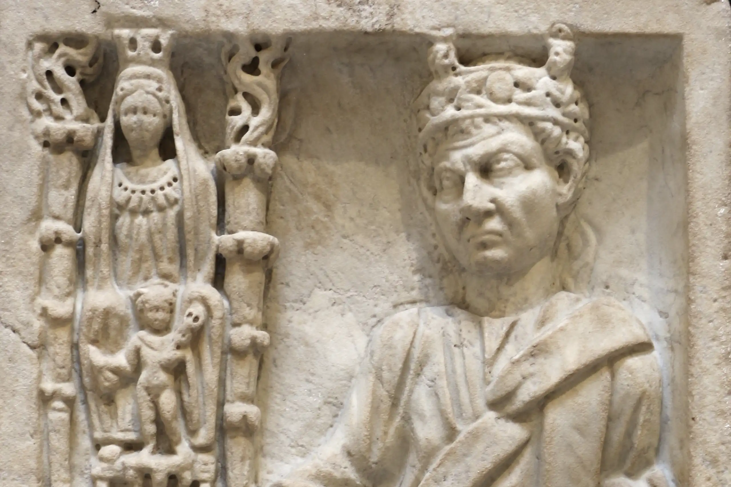 A marble relief with several figures engraved on it: on the side is a column with an adorned woman with another companion. Next to them is a man with a crown, with one hand on a pedestal.