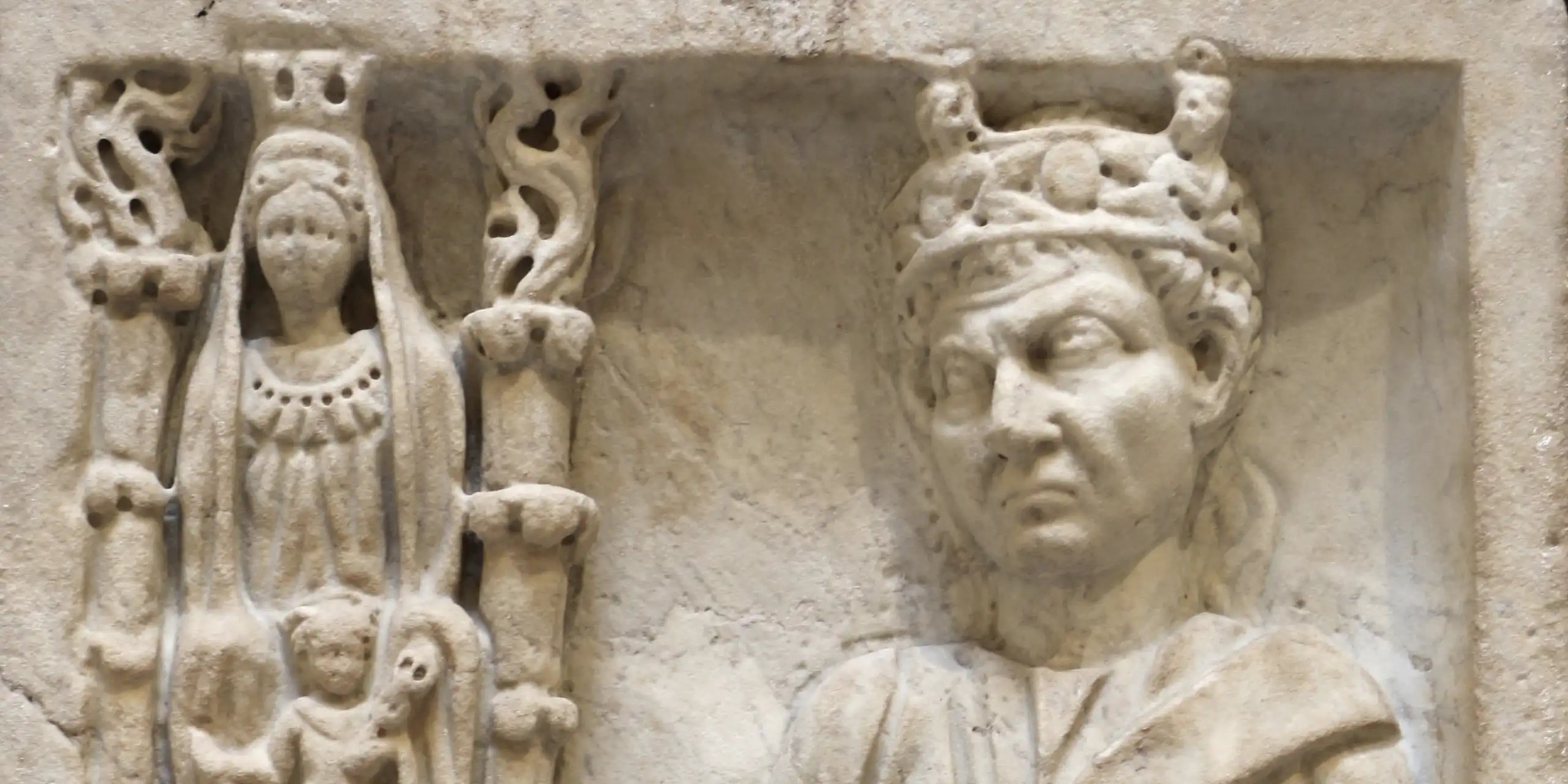 A marble relief with several figures engraved on it: on the side is a column with an adorned woman with another companion. Next to them is a man with a crown, with one hand on a pedestal.