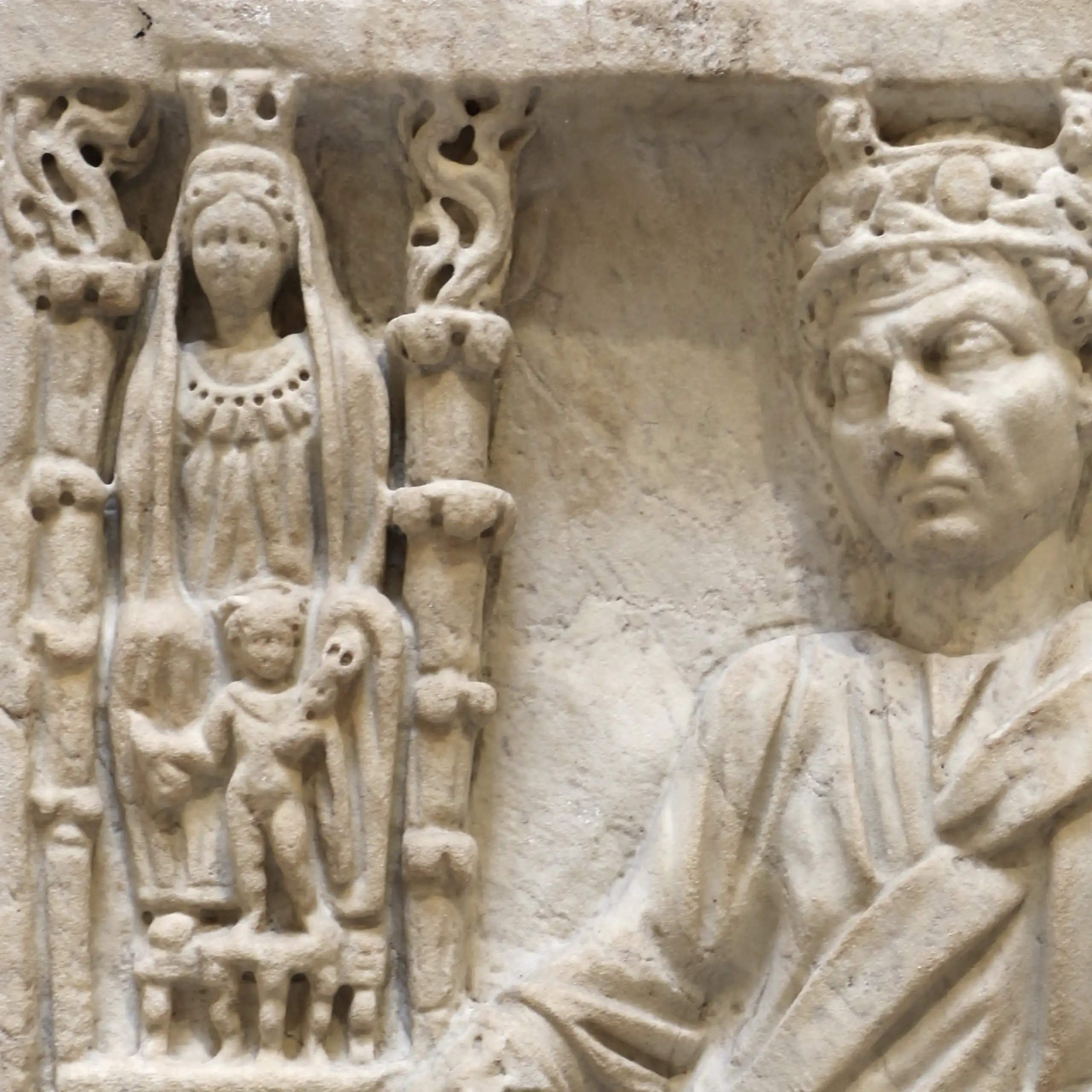A marble relief with several figures engraved on it: on the side is a column with an adorned woman with another companion. Next to them is a man with a crown, with one hand on a pedestal.