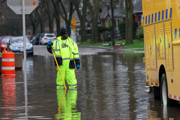 A city worker in reflective clothes stands knee-deep in a flooded street.