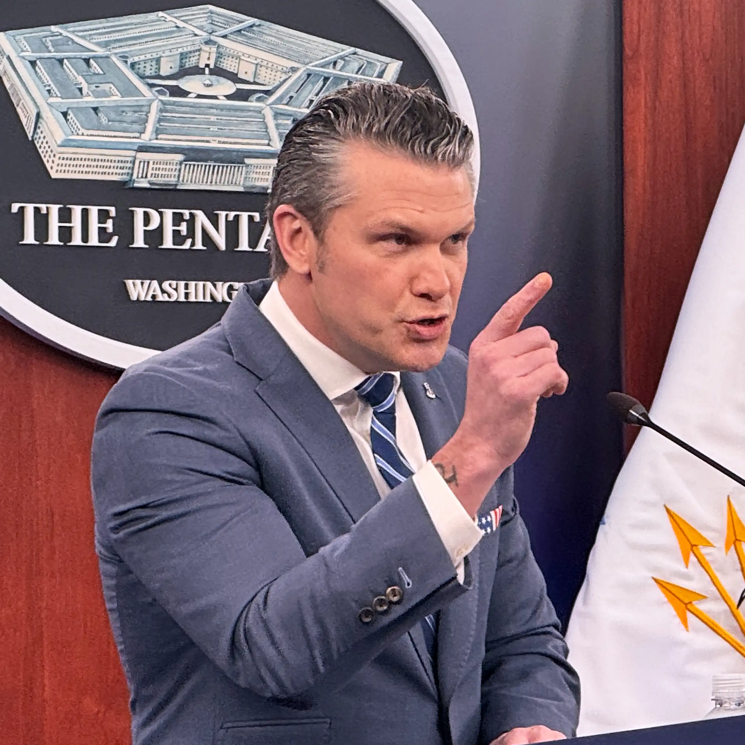 A man glowers with one hand raised as he stands behind a podium at a press briefing.