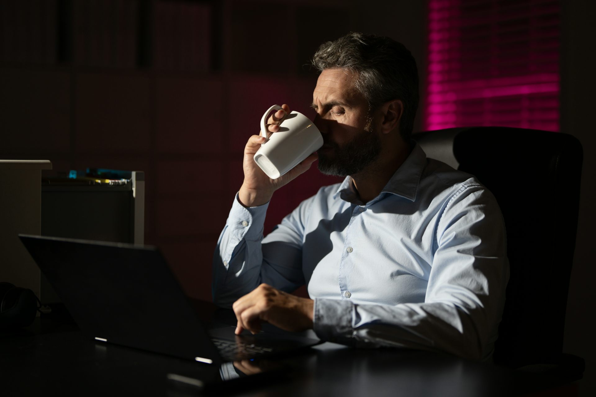Un homme au bureau est plongé dans le noir et boit une boisson chaude.