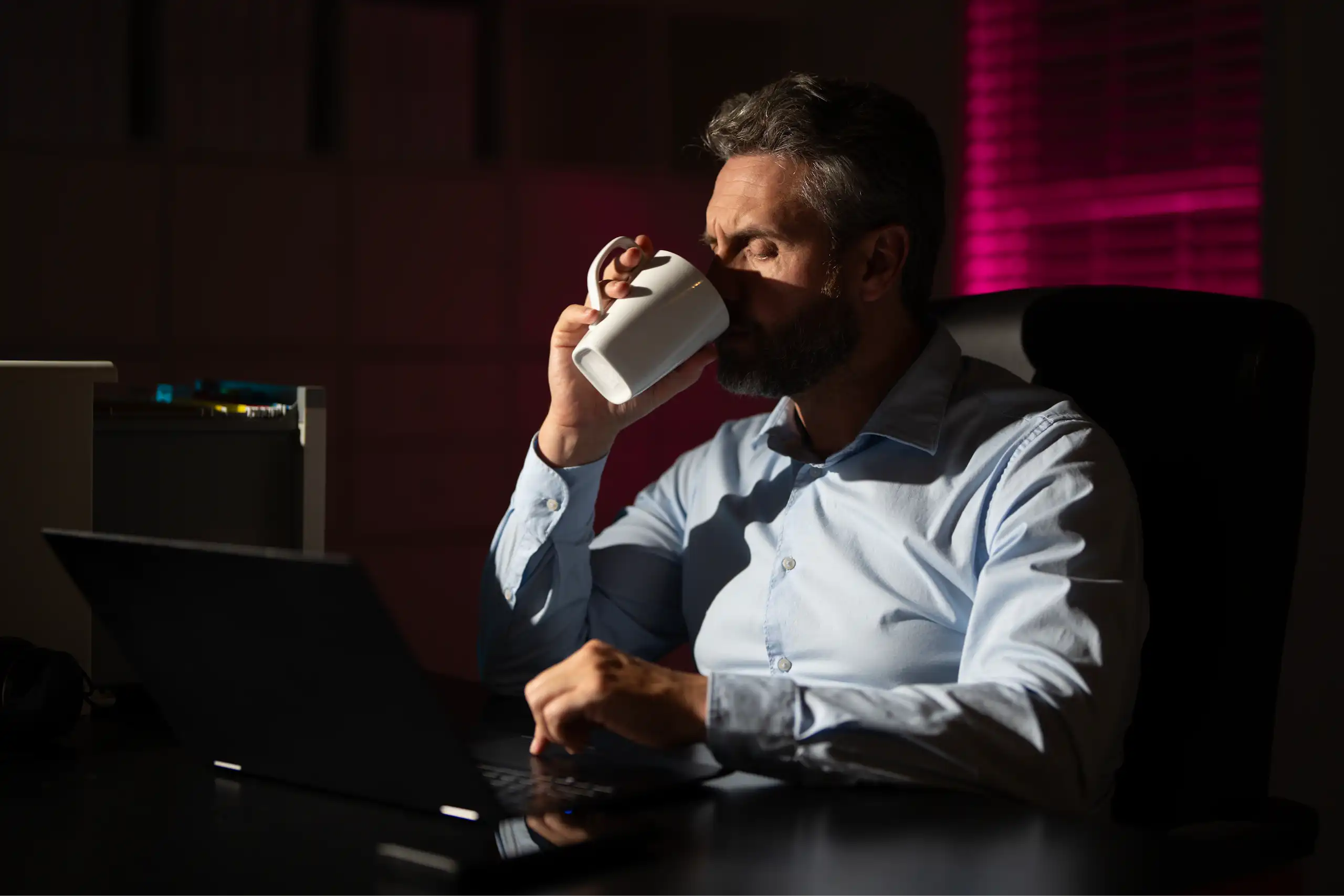 Un homme au bureau est plongé dans le noir et boit une boisson chaude.