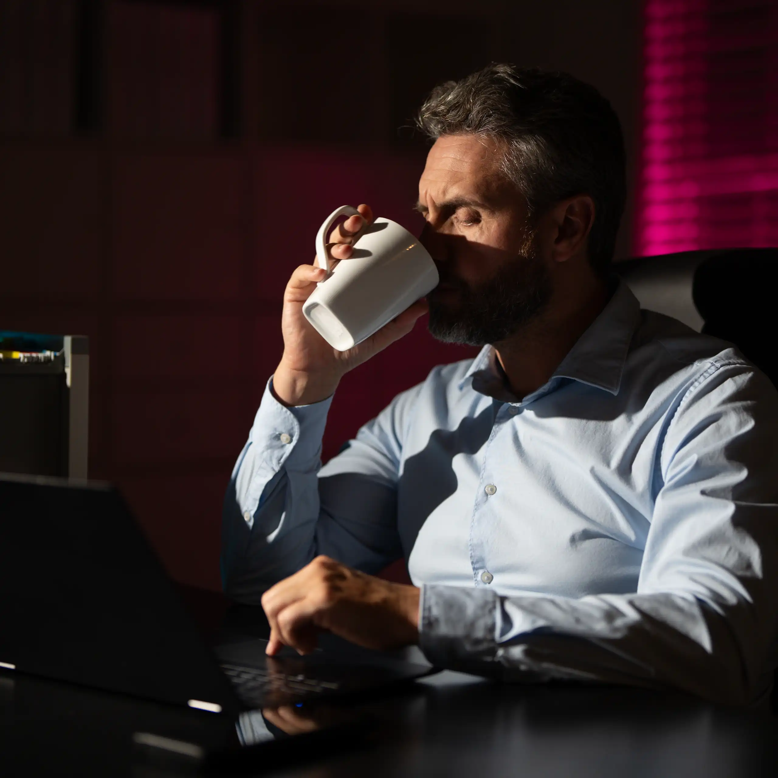 Un homme au bureau est plongé dans le noir et boit une boisson chaude.