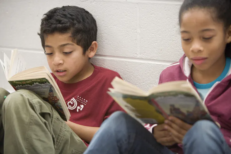 Two children who look about eight or nine years old sit side by side against a white wall and read books.