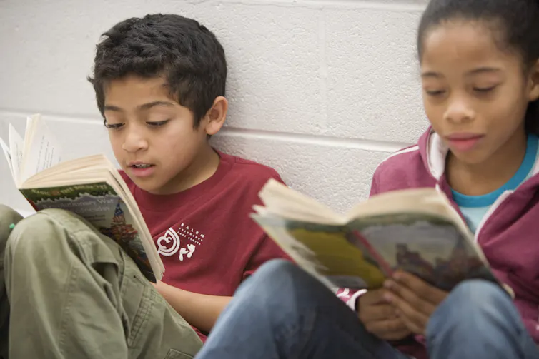 Two children who look about eight or nine years old sit side by side against a white wall and read books.