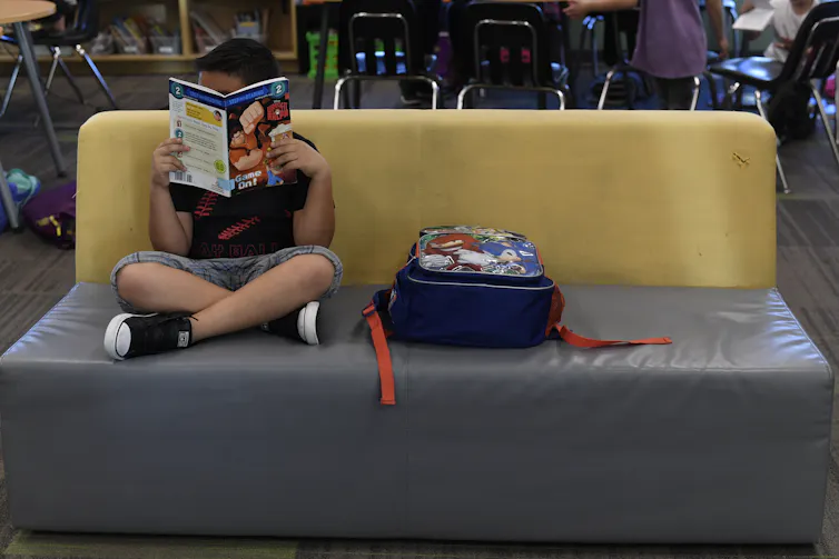 A child sits on a couch with his legs crossed, his face obstructed by a book. He sits next to a backpack.