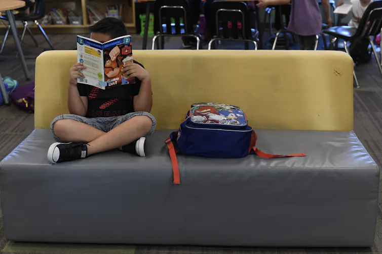 A child sits on a couch with his legs crossed, his face obstructed by a book. He sits next to a backpack.