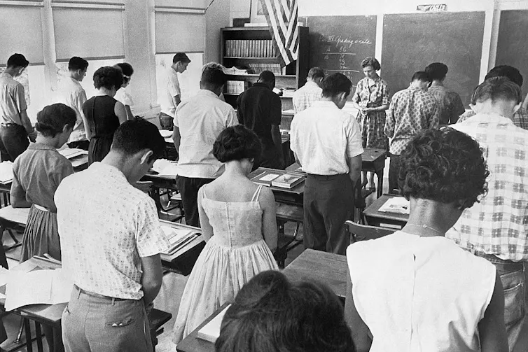 A black and white photograph, taken from the back of a classroom, shows a few rows of students standing with their heads bowed.