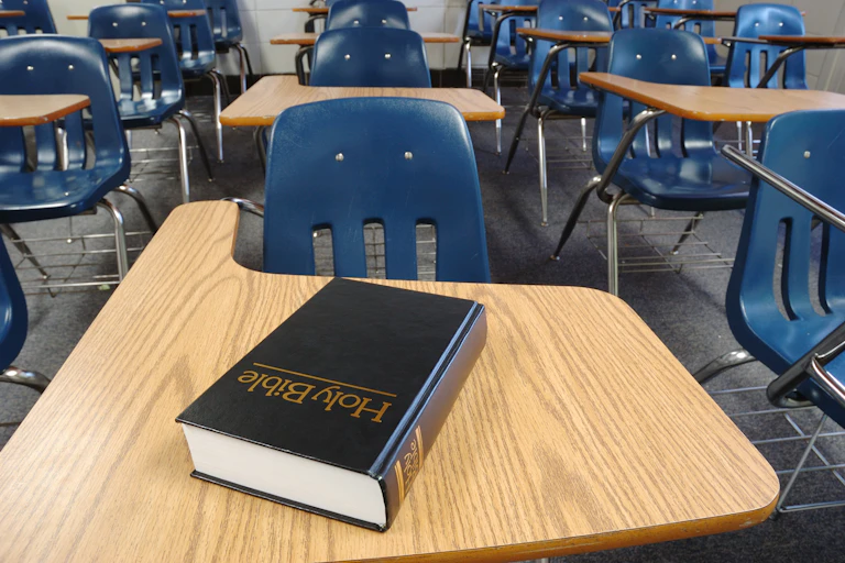 A book that says 'Holy Bible' on the cover rests on the top of a desk in a room full of desks attached to blue plastic chairs.