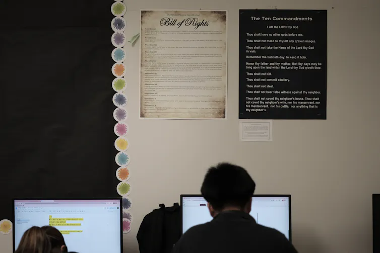 Two students, facing away from the camera, read text on computers positioned up against a white wall.