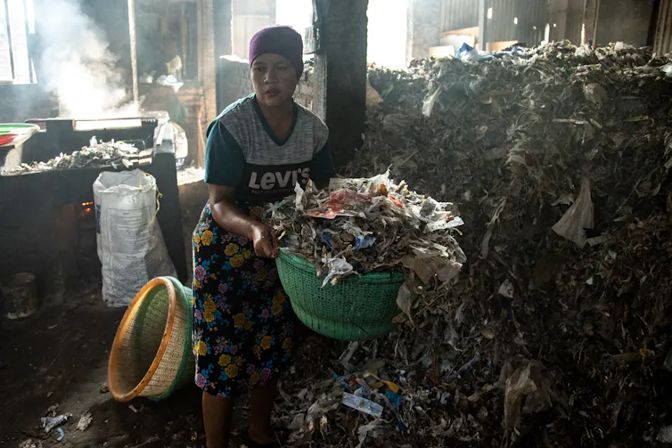 A woman carries a basket with plastic waste from a large pile in a room.