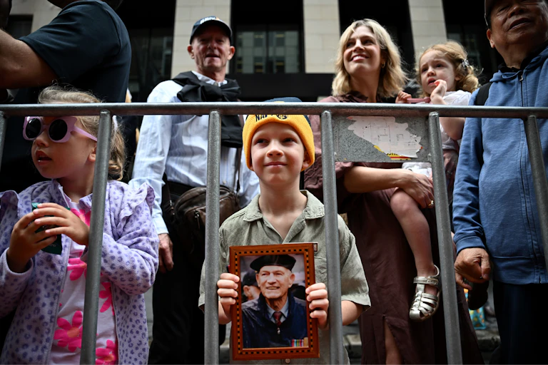 A young boy holds the photo of a veteran, while standing in a crowd. Another young child stands behind him. A woman holds a small child on her hip behind.