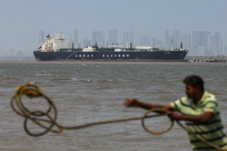 An Indian port worker throws rope over a stanchion as a tanker arrives in Mumbai port in the background.
