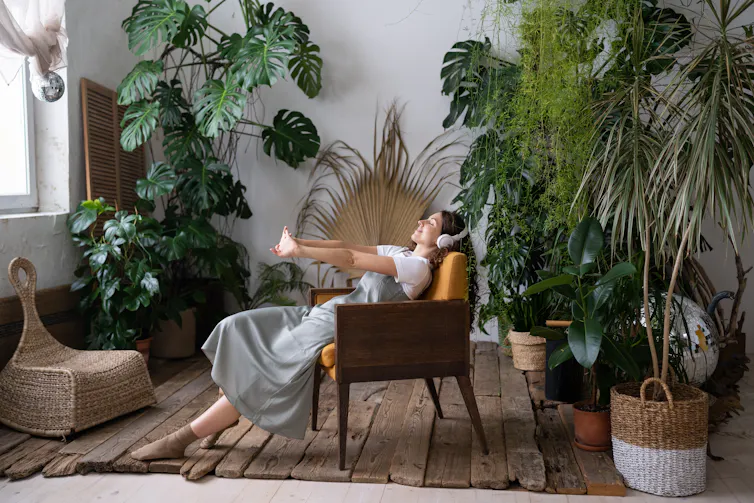 Woman wearing headphones leaning back in armchair, surrounded by large houseplants