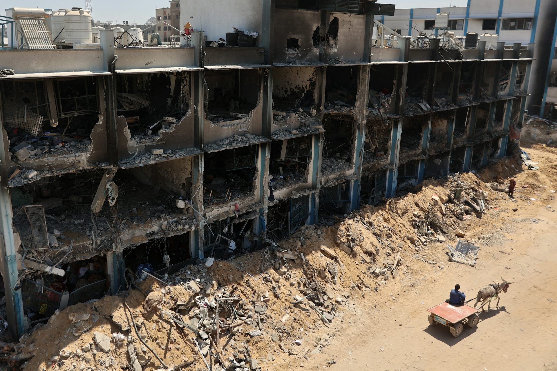 A destroyed building is seen with a person on a cart pulled by a donkey nearby.