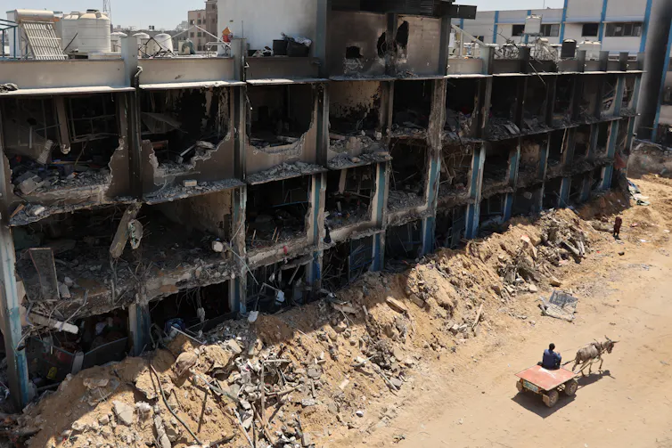 A destroyed building is seen with a person on a cart pulled by a donkey nearby.