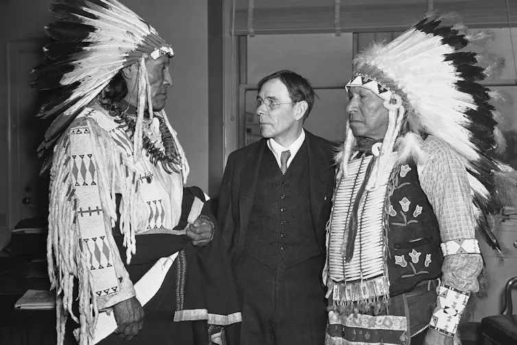 'Bombardeando nuestros pequeños corazones': cómo Trump se está aprovechando del duradero apetito de destrucción de Estados Unidos 1 Dos hombres nativos con atuendos tribales están a cada lado del hombre blanco.