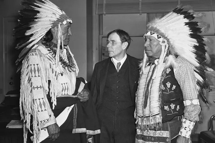 Two Indigenous men in tribal regalia stand on either side of a white man.