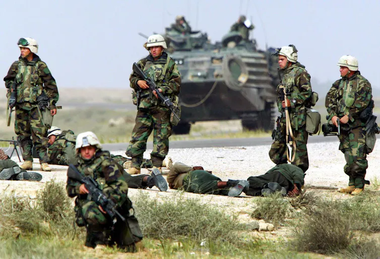 Heavily armed soldiers stand around men lying on the ground. A tank is behind them.