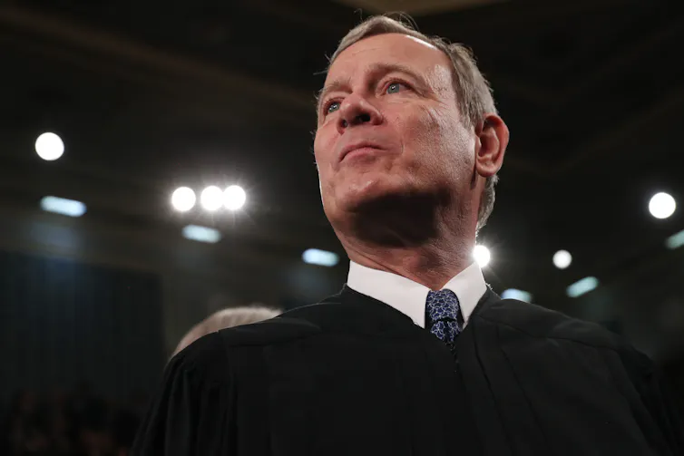 A man with short hair, wearing a black robe over a white shirt and blue tie.